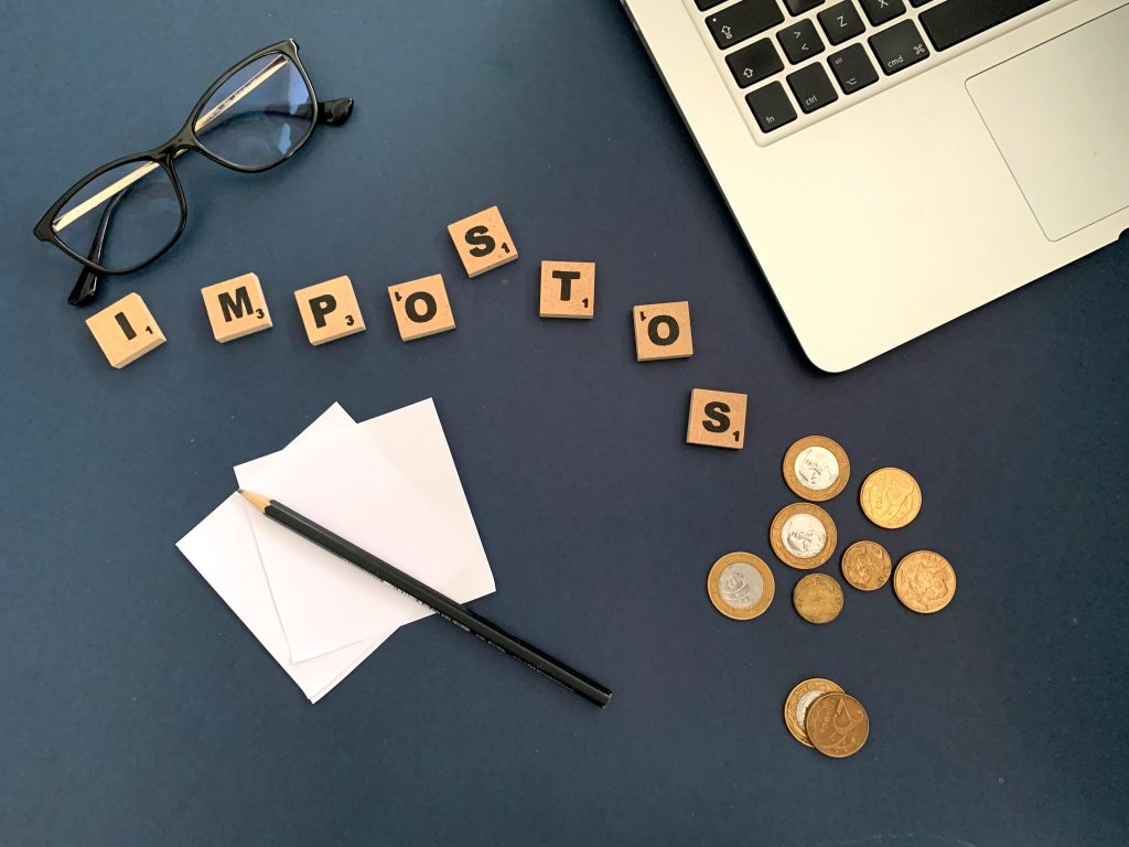Top view of a workspace with coins, glasses, and the word 'impostos' spelled out, symbolizing taxes.